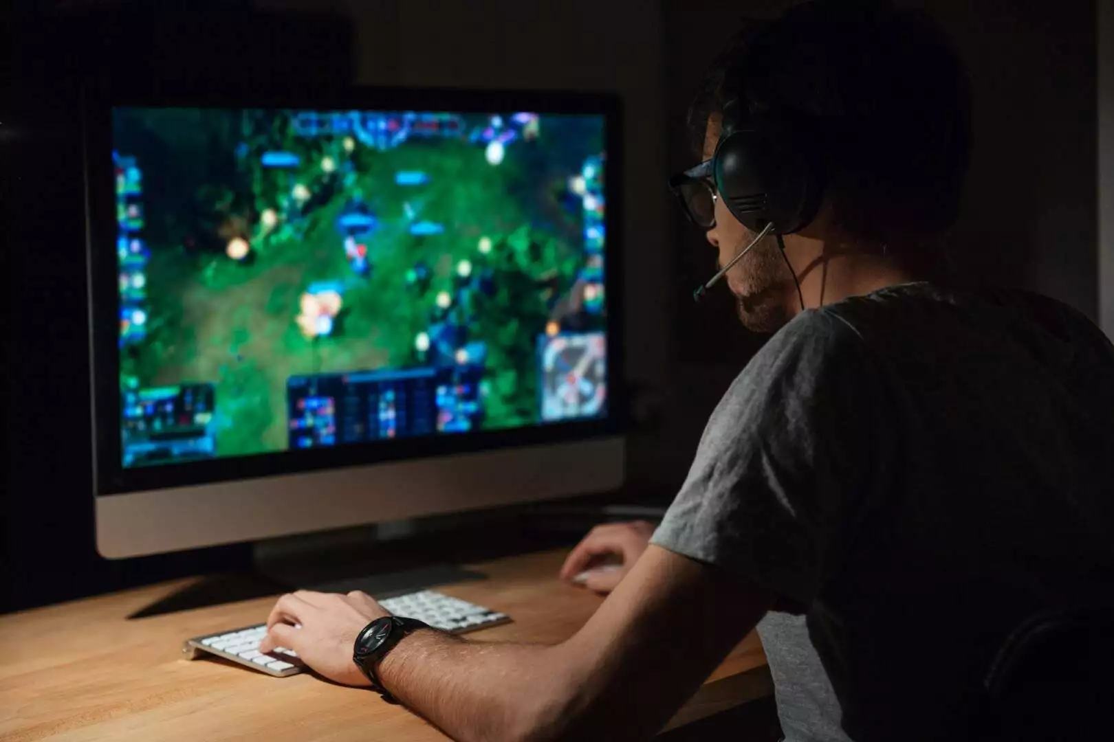 Back view of concentrated young gamer in headphones and glasses using computer for playing game at home.